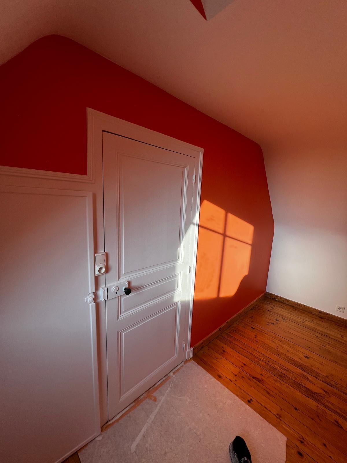 Mur rouge vif sous une pente de toit, avec une porte et des plinthes peintes en blanc